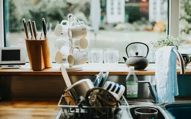 Bancada de cozinha ao lado da janela, com escorredor cheio, canecas e bule depois do uso do dia