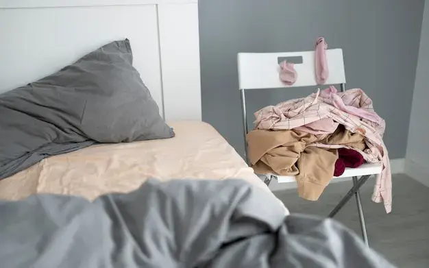 Cadeira de quarto com roupas acumuladas no assento, ao lado da cama desfeita