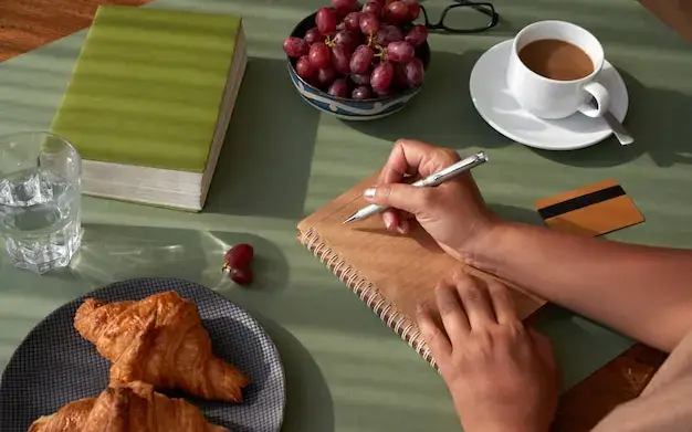 Mãos anotando num caderno sobre a mesa, ao lado de uma xícara de café e do prato do café da manhã