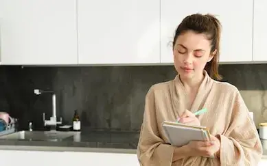 Mulher em pé na cozinha escrevendo a lista de compras num caderno apoiado na bancada
